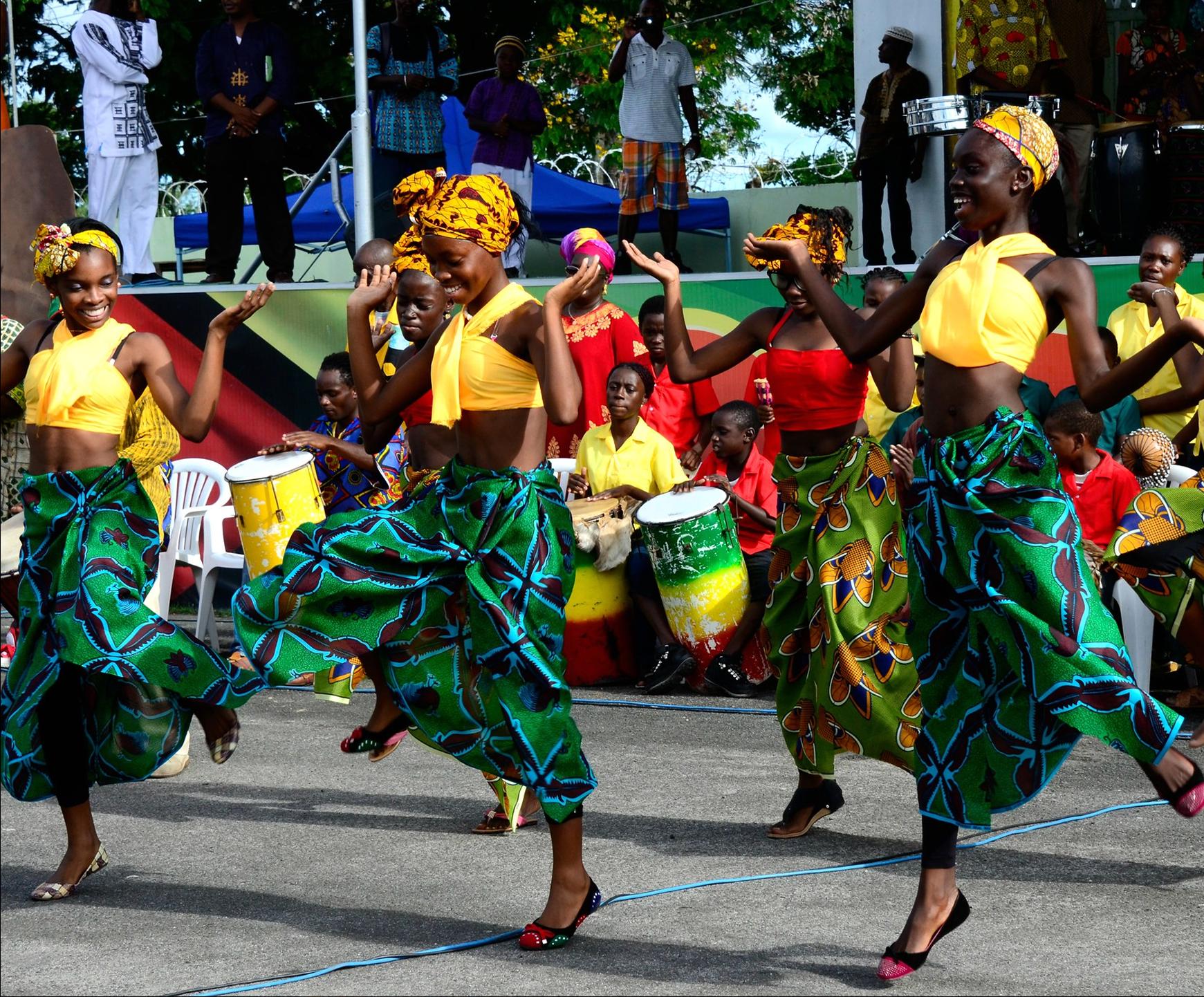 The image depicts a group of people dancing in a vibrant outdoor setting, with a stage visible in the background. The dancers are dressed in various colorful outfits, including green patterned skirts and yellow tops or headwraps, as well as red dresses or tops.